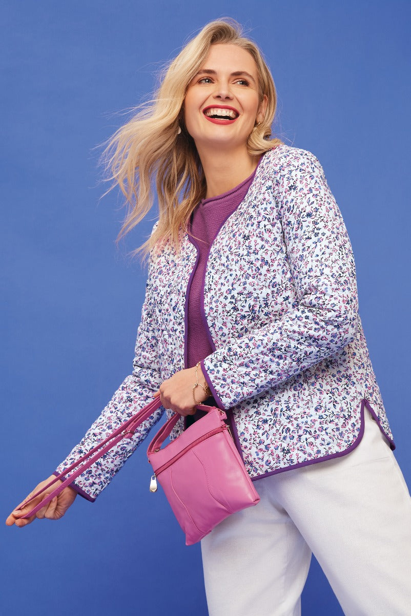 Lily Ella Collection floral print jacket in white and purple, stylish woman wearing purple sweater, white trousers, and pink handbag, trendy spring outfit, vibrant blue background
