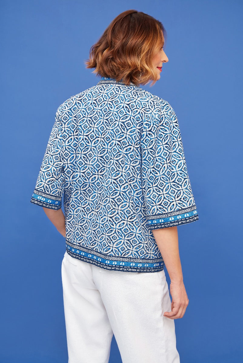 Lily Ella Collection blue and white patterned blouse with relaxed fit and short sleeves, paired with white trousers for a casual chic look, on a blue background.