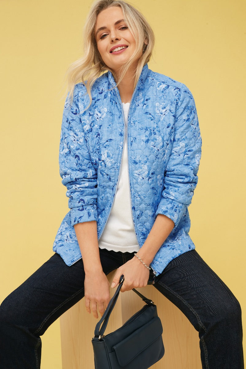 Lily Ella Collection women's blue floral quilted jacket, stylish patterned lightweight coat, casual chic outfit with denim jeans and white top, accessorized with navy handbag on a yellow background