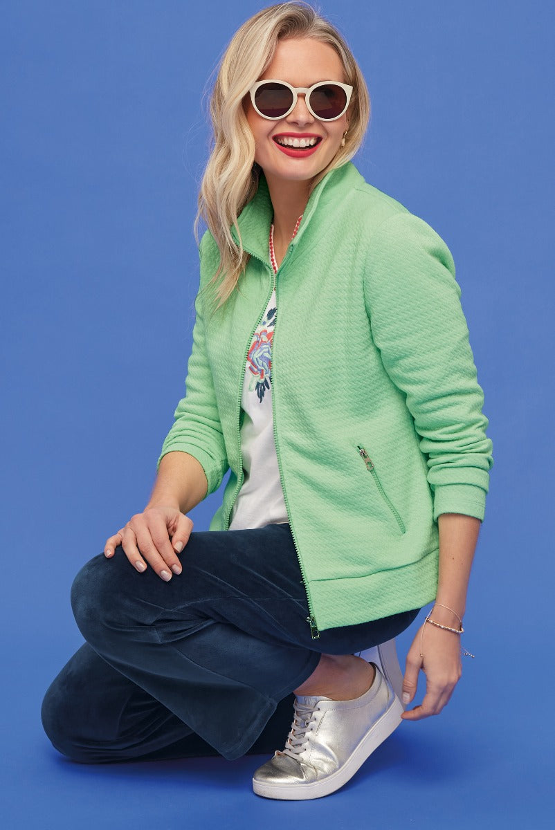 Lily Ella Collection vibrant green quilted zip-up jacket, styled with white embroidered blouse, navy trousers, metallic silver shoes, and white round sunglasses for a casual chic look.