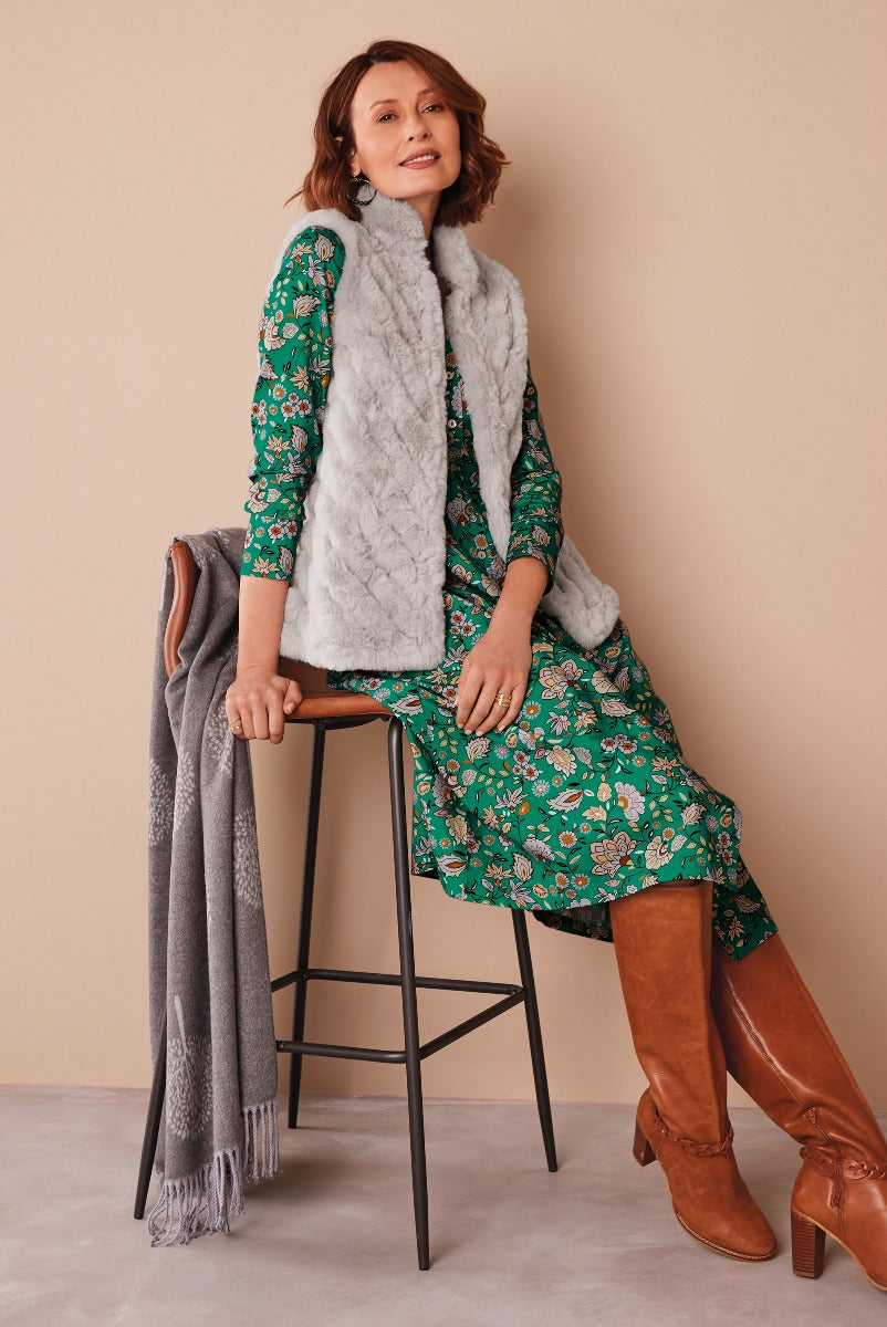 Lily Ella Collection stylish woman in green floral dress with gray faux fur vest and tan knee-high boots, accessorized with a draped gray fringed scarf over a black bar chair.