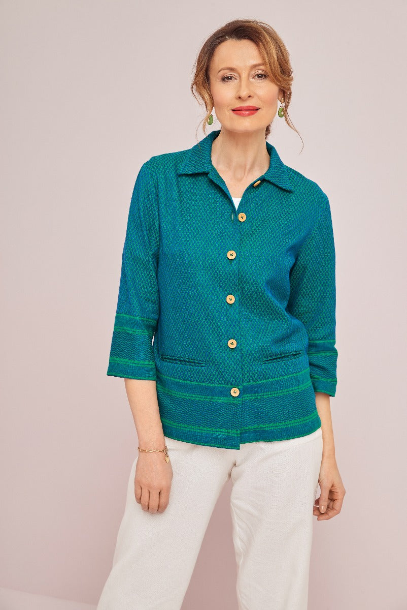 Lily Ella Collection teal green textured jacket with golden buttons, paired with white trousers, presented by a model against a pink background.