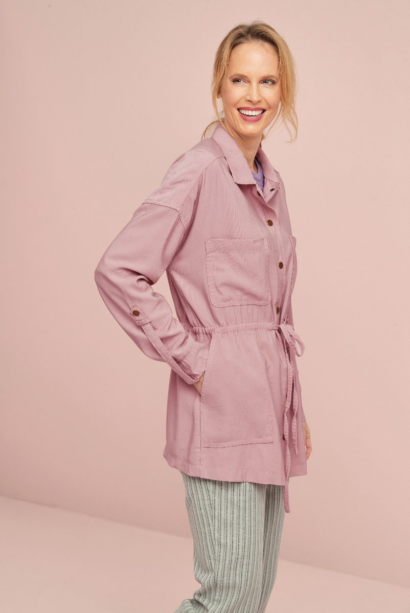 Smiling woman modeling Lily Ella Collection pink corduroy shirt jacket with tie waist paired with casual striped trousers against a pink background