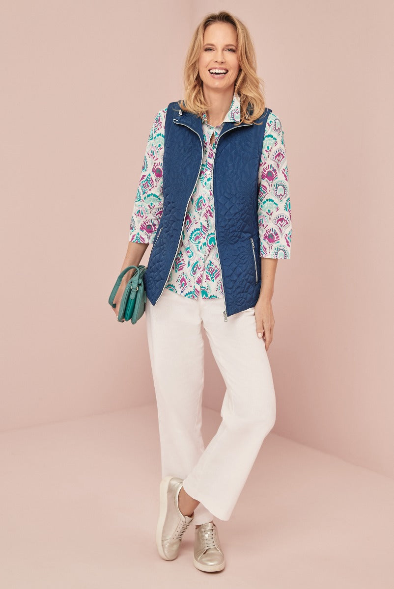 Lily Ella Collection navy quilted gilet paired with white paisley pattern blouse and white casual trousers, woman smiling in a trendy spring outfit with metallic sneakers and teal handbag.
