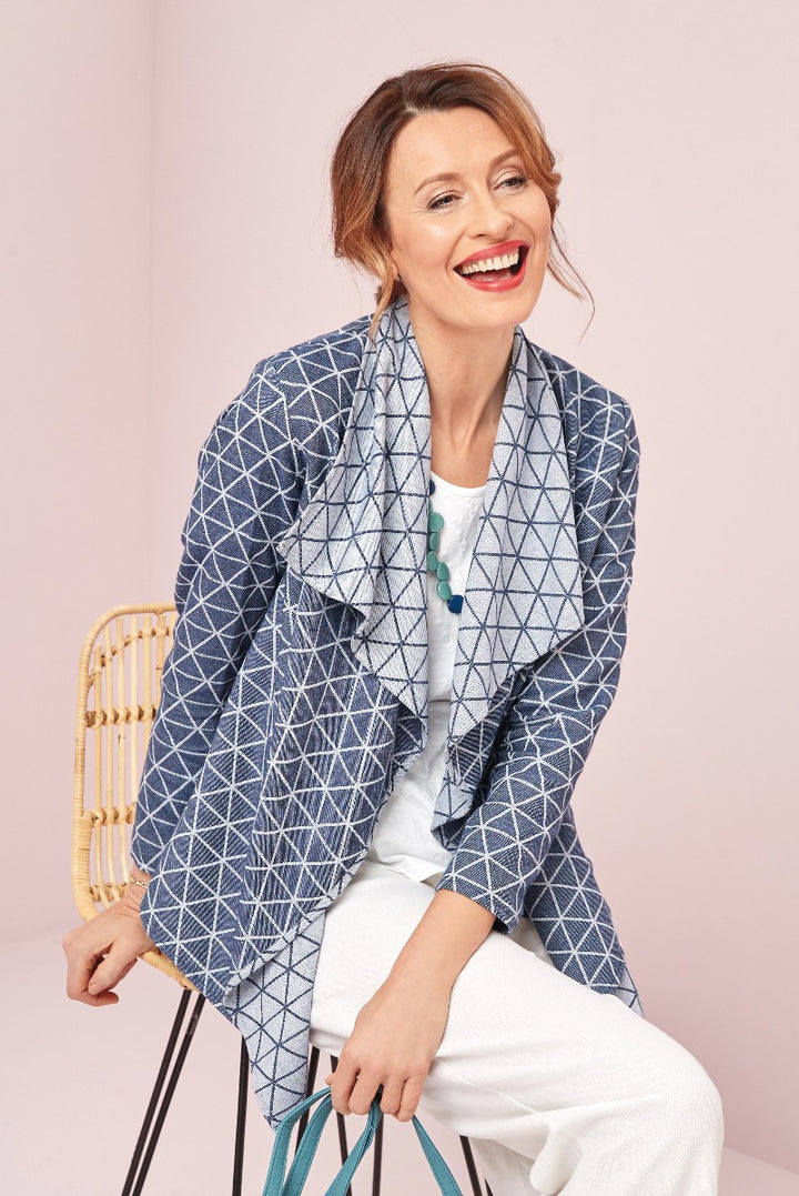 Lily Ella Collection cheerful woman modeling blue geometric-patterned blazer with white pants and statement necklace sitting on a metal chair against a pink background