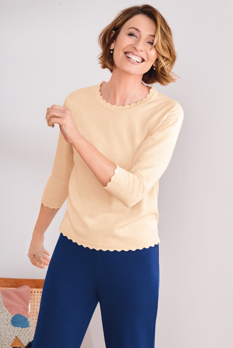 Lily Ella Collection stylish woman in a pale yellow scalloped-edge sweater with comfortable blue trousers smiling in a bright indoor setting, showcasing casual elegance