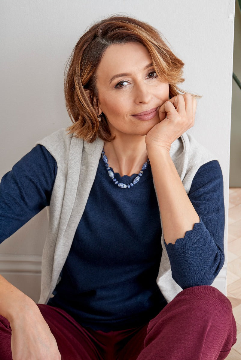 Lily Ella Collection stylish woman in layered look, featuring a navy blue top and light grey cardigan, accessorized with a chunky blue necklace, paired with maroon trousers, casual chic ensemble for mature women.