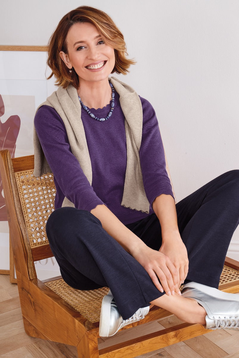 Smiling woman sitting in a wicker chair wearing Lily Ella Collection's purple sweater, beige cardigan, and navy trousers, accessorized with a blue necklace and paired with silver sneakers.