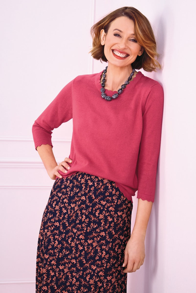 Lily Ella Collection elegant woman in a raspberry 3/4 sleeve knit jumper and floral pattern navy skirt with statement necklace smiling against a pink background