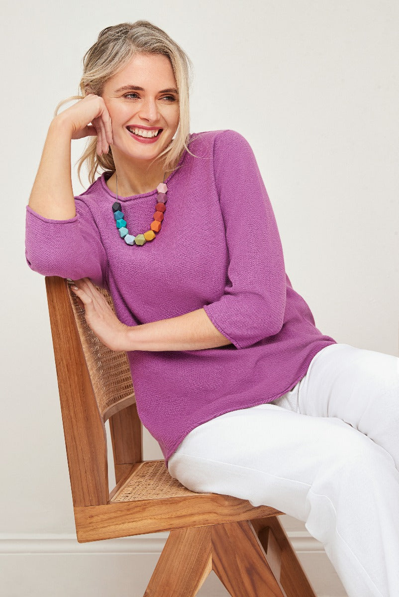 Lily Ella Collection purple knit jumper, stylish women's 3/4 sleeve top, white trousers, model posing on a wooden chair, chic comfortable fashion, colorful statement necklace accessory.