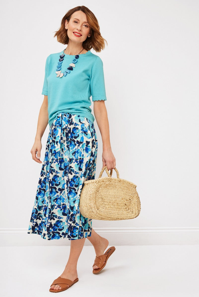 Lily Ella Collection teal short-sleeve top and blue floral print midi skirt, woman carrying straw tote bag and wearing brown sandals.