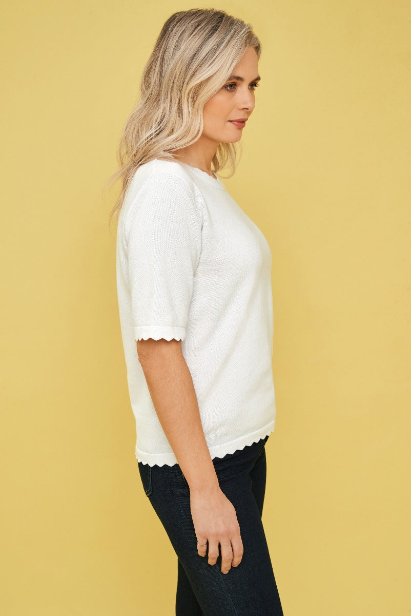 Lily Ella Collection white scallop-trimmed knit top styled with classic denim, women's casual elegant clothing, model side view against a yellow background