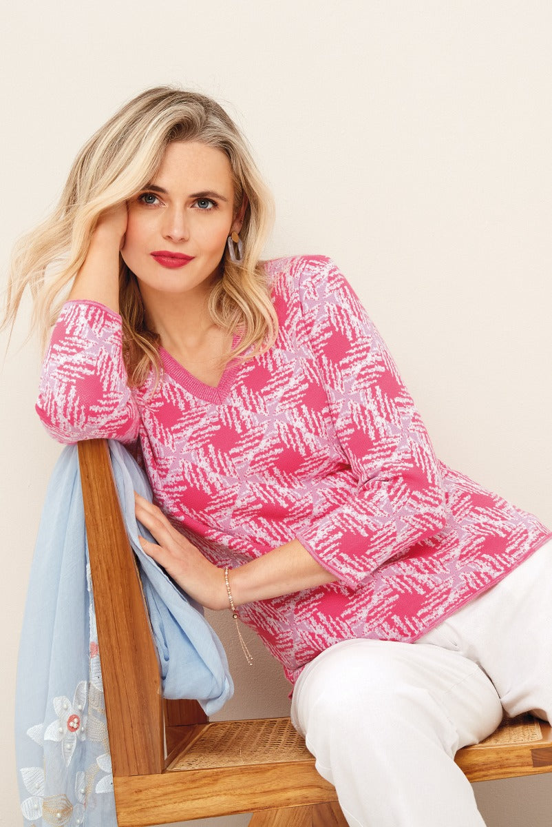 Lily Ella Collection pink and white patterned sweater, stylish v-neck knitwear, elegant casual look, paired with white trousers and blue scarf accessory.