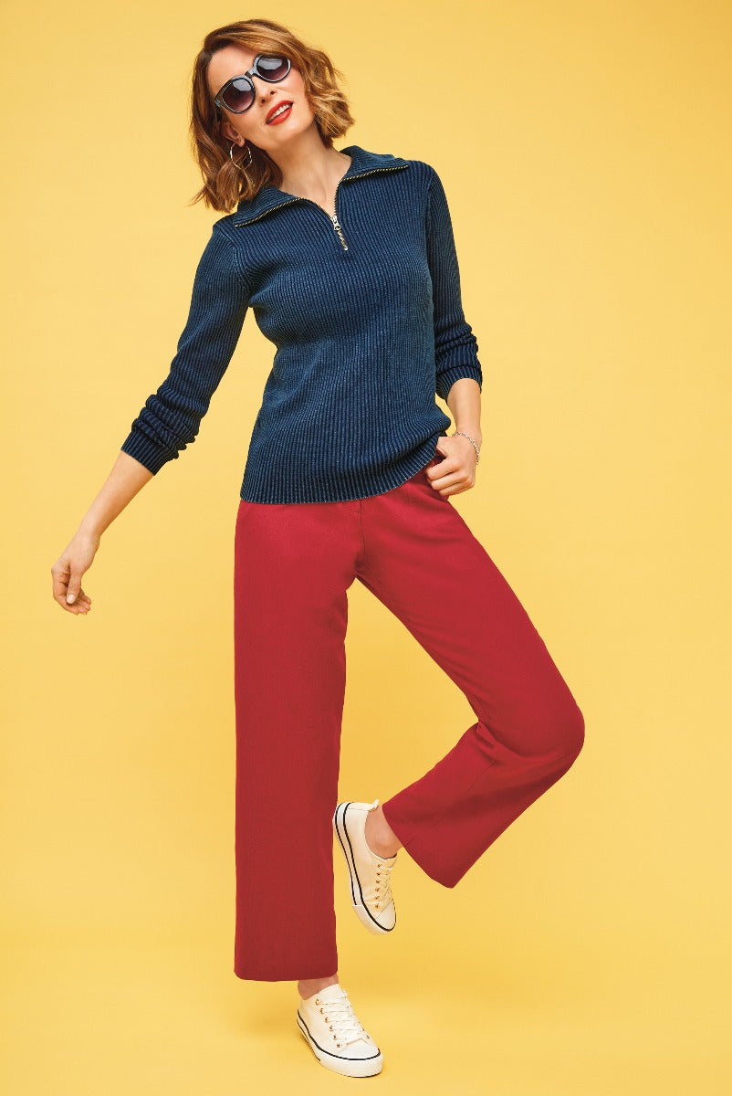 Lily Ella Collection stylish woman wearing navy blue ribbed zip neck jumper and red wide-leg trousers with white sneakers on a yellow background.