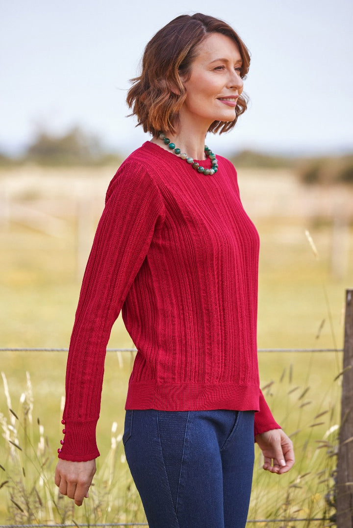 Lily Ella Collection elegant red knit sweater with button sleeve detail, styled with blue jeans and a green beaded necklace, casual chic women's fashion in outdoor setting.