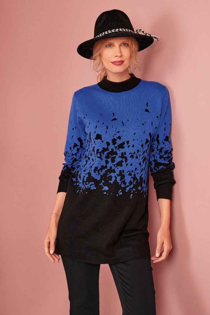 Lily Ella Collection model wearing a blue and black gradient splatter pattern sweater with black turtleneck and pants, accented with a chic black hat with houndstooth ribbon.