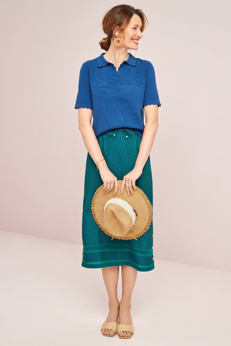 Lily Ella Collection elegant blue scallop-trimmed blouse and emerald green patterned midi skirt, accessorized with straw sun hat and beige sandals, sophisticated summer outfit idea.