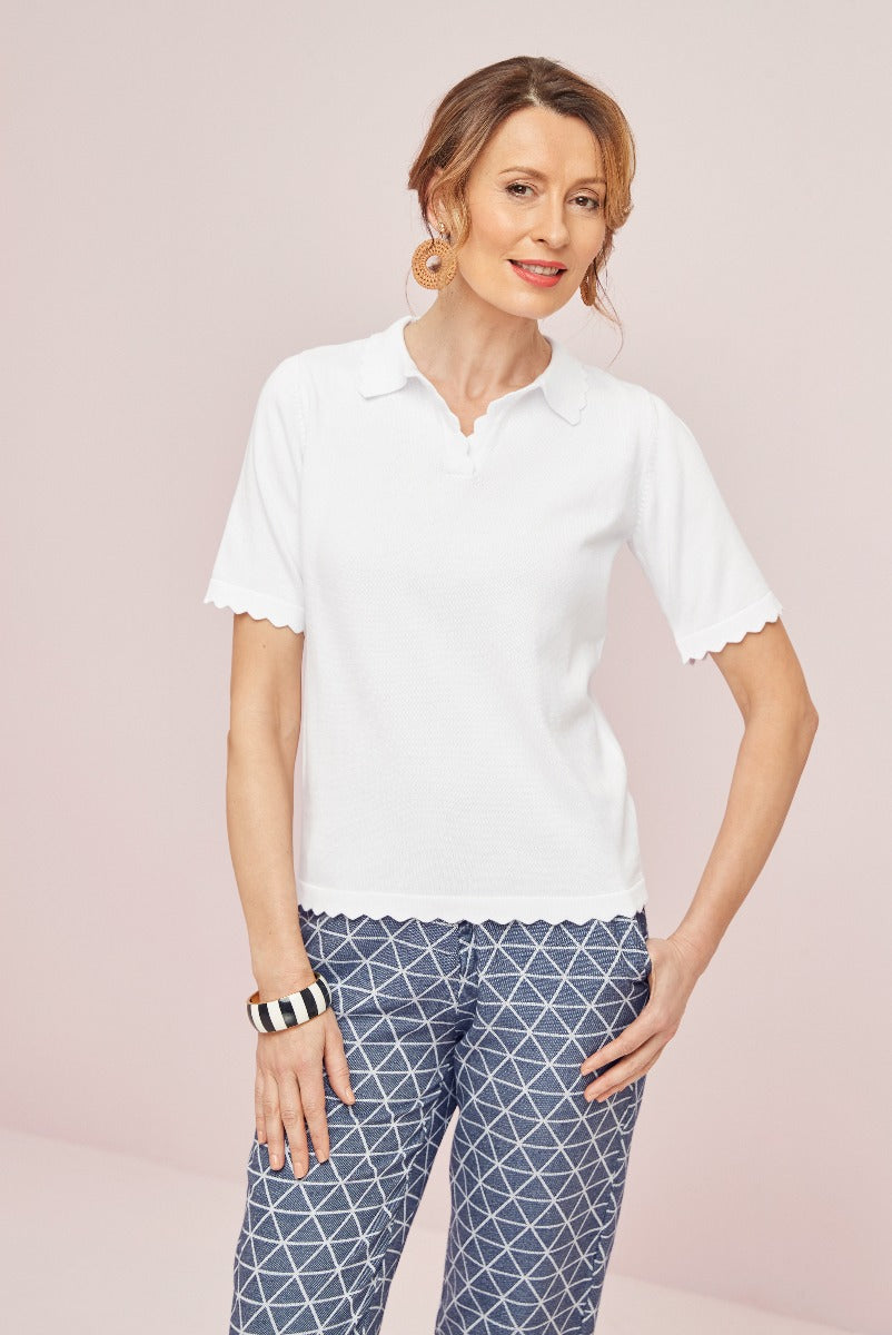 Lily Ella Collection white textured polo shirt with scalloped collar and cuffs paired with geometric patterned navy trousers, styled with hoop earrings and a striped bangle on a soft pink background.