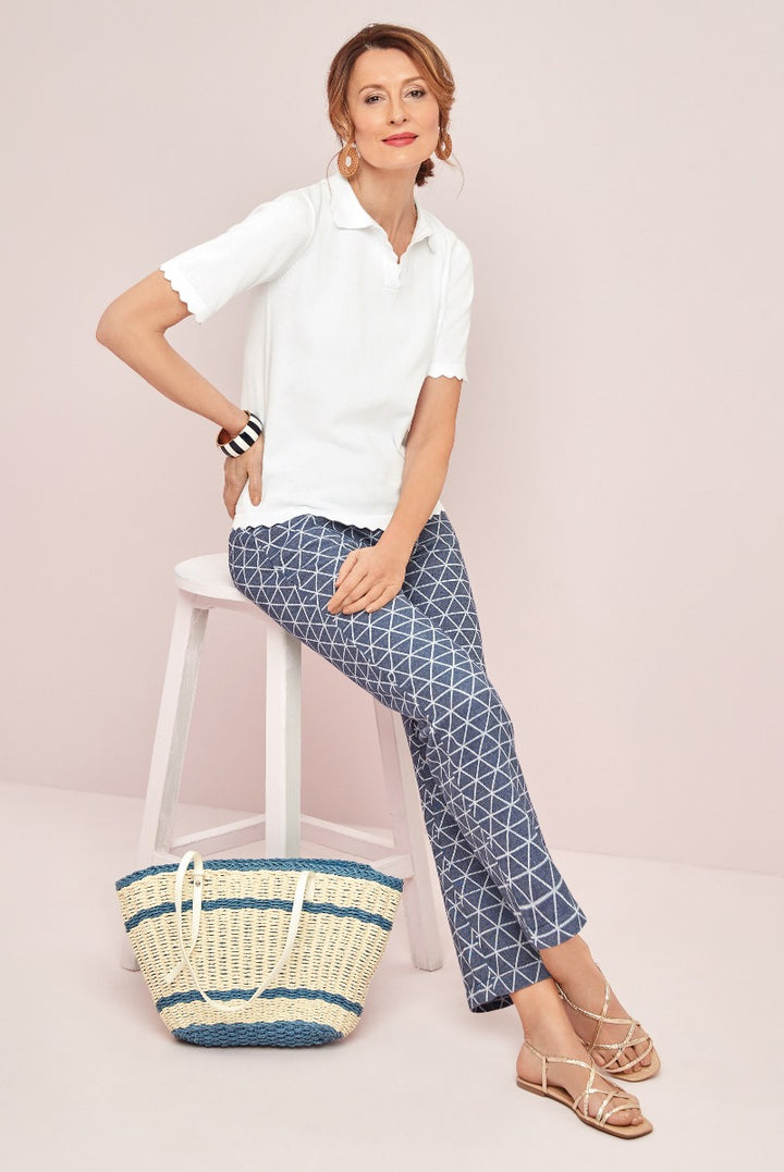 Lily Ella Collection casual summer outfit featuring white scallop-edged polo shirt, geometric pattern navy blue cropped trousers, and a striped yellow and blue straw tote bag with stylish strappy sandals.