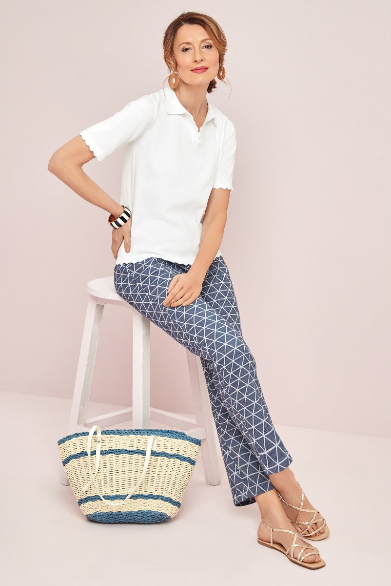 Lily Ella Collection stylish casual women's outfit with white scalloped edge polo shirt, geometric patterned blue ankle length trousers, coordinated straw basket bag, and metallic strappy sandals.