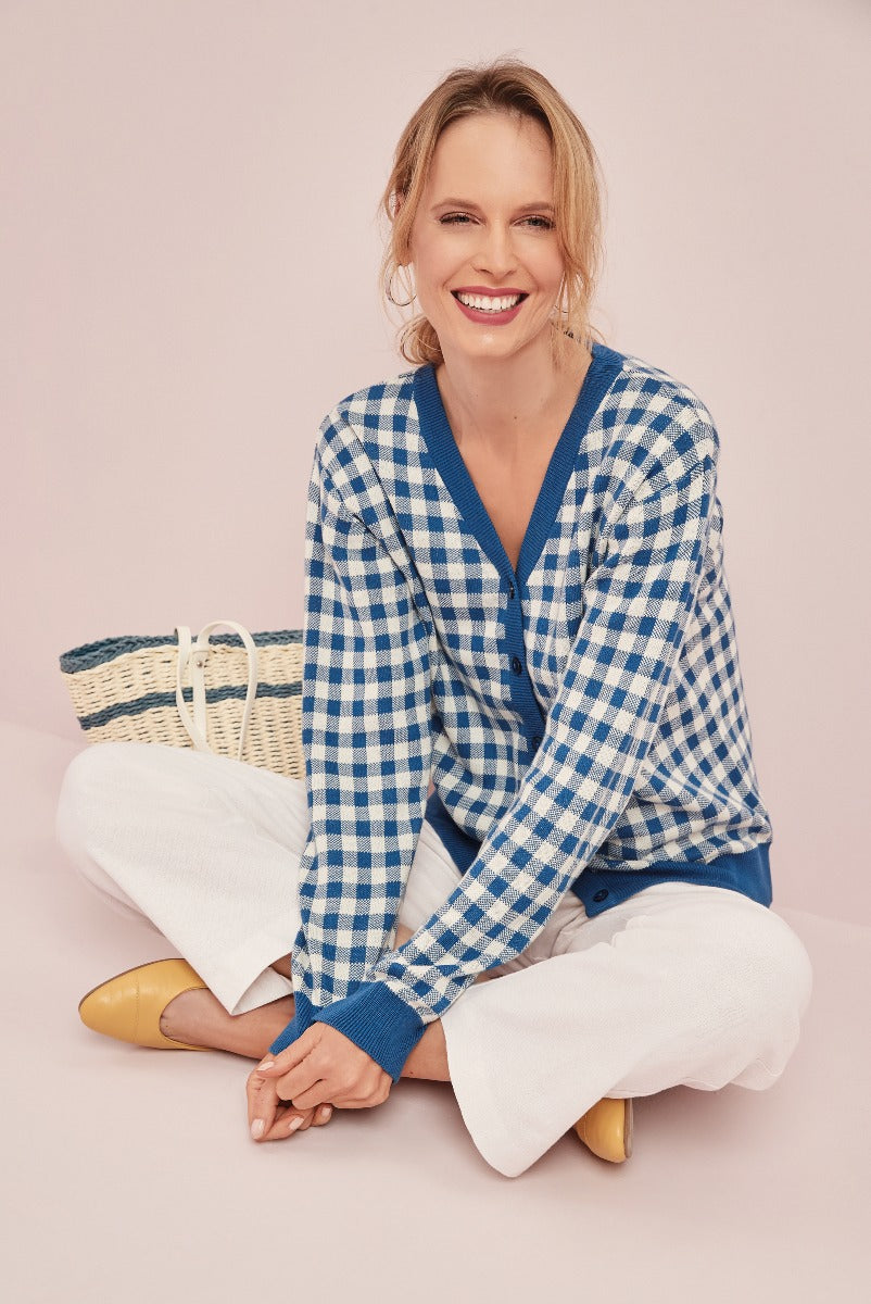 Lily Ella Collection model in a blue and white checkered cardigan, white trousers, and mustard yellow mules, sitting with a straw handbag against a pastel pink background.