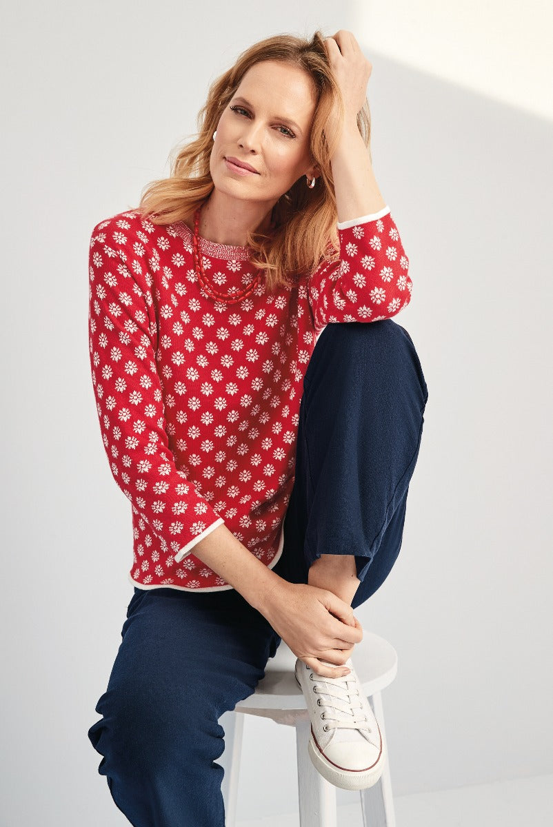 Lily Ella Collection red patterned sweater with white trim paired with navy casual trousers and white sneakers on seated model