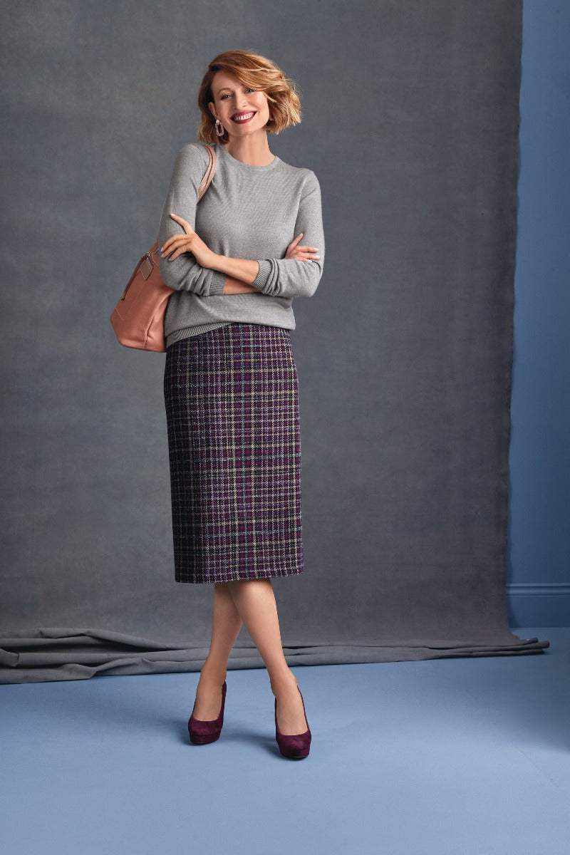 Lily Ella Collection fashion model wearing a casual grey sweater, trendy purple checkered pencil skirt, matching purple heels, and a stylish blush handbag, posing with confidence on a blue-grey backdrop.