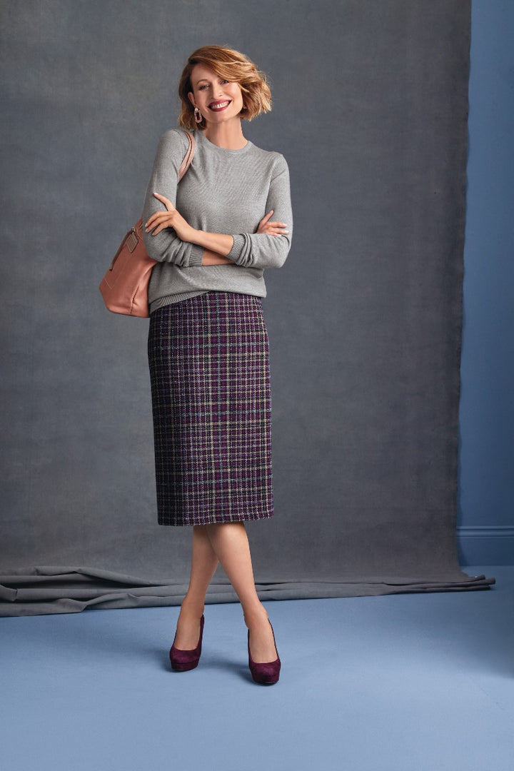 Lily Ella Collection stylish woman posing in a chic grey sweater and plaid purple pencil skirt with matching velvet heels and a tan shoulder bag on a grey backdrop