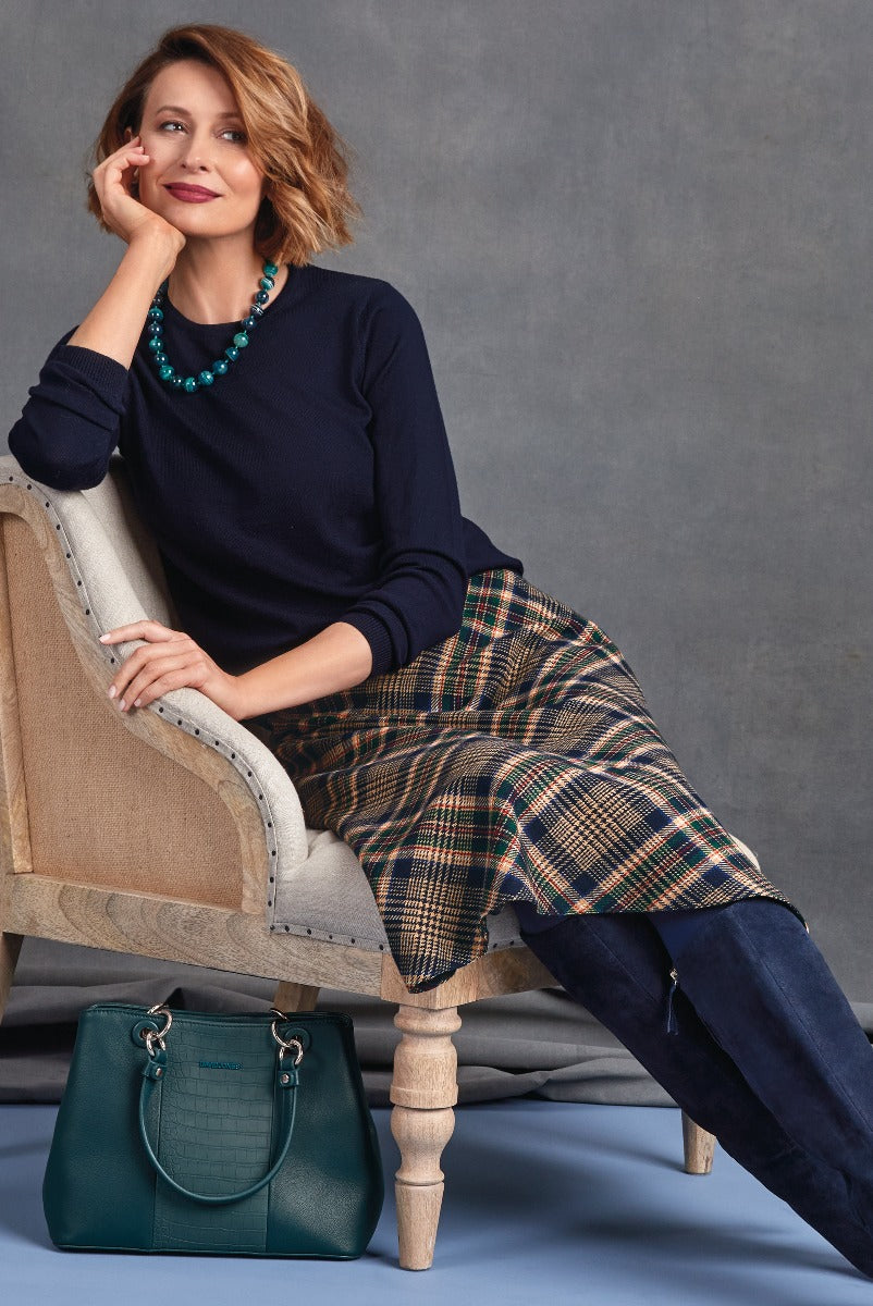 Lily Ella Collection elegant navy blue sweater with plaid skirt, stylish green handbag, and statement necklace, woman posing fashionably on upholstered chair.