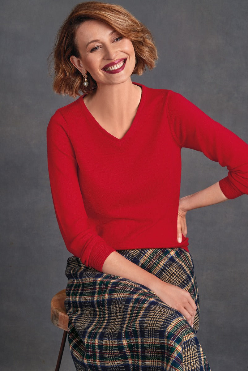 Lily Ella Collection vibrant red V-neck sweater paired with stylish tartan skirt, smiling female model posing fashionably on stool.