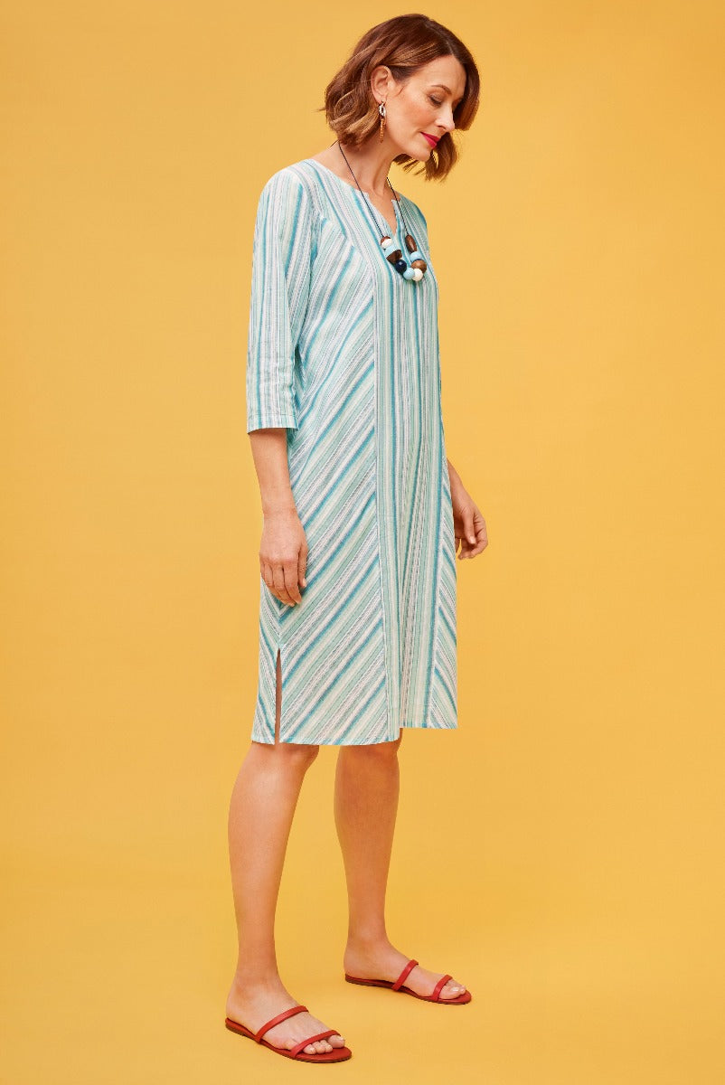 Lily Ella Collection aqua blue and white striped midi dress with elbow-length sleeves, V-neck, and front pockets on a model against a yellow background, paired with red sandals and statement necklace.