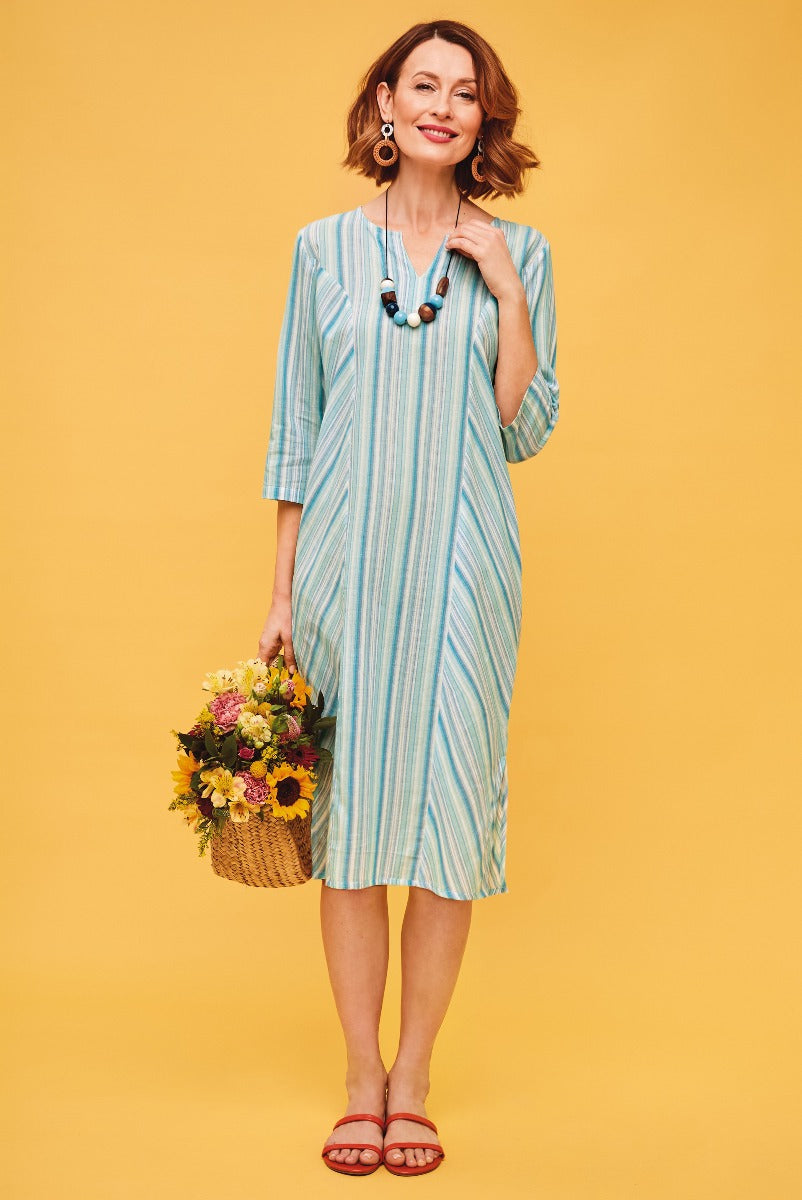 Lily Ella Collection woman wearing blue and white striped midi dress with three-quarter sleeves and red sandals, holding a basket of flowers against a yellow background.