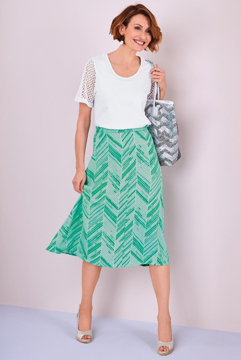 Lily Ella Collection summer fashion, smiling woman wearing a white eyelet blouse paired with a green leaf-patterned midi skirt, accessorized with a silver patterned tote bag and cream open-toe heels.