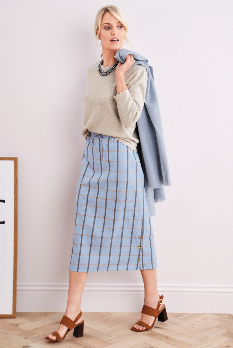Lily Ella Collection elegant woman in beige crew neck jumper and blue checkered midi skirt with side button detail, holding a light blue coat, accessorized with layered necklaces, paired with brown block heel sandals.