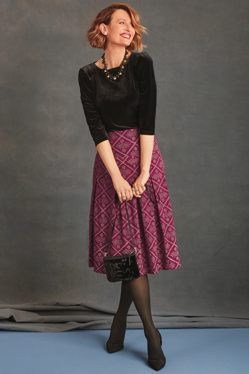 Lily Ella Collection stylish woman posing in a black velvet top and vibrant purple patterned midi skirt, accessorized with black statement necklace and clutch, on a gray backdrop.