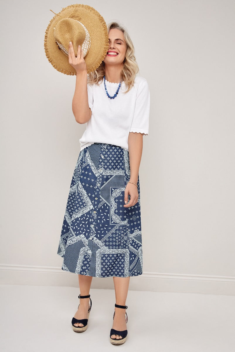 Lily Ella Collection model smiling and holding a straw hat wearing a white scallop trim top and a blue patterned mid-length skirt with navy wedge sandals.