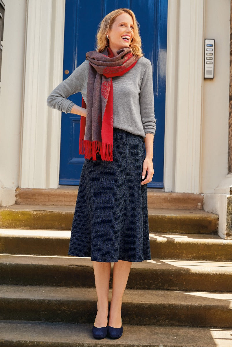 Lily Ella Collection fashion model wearing a grey sweater, red fringed scarf, and navy blue pleated midi skirt, smiling and standing on outdoor steps, stylish women's autumn clothing