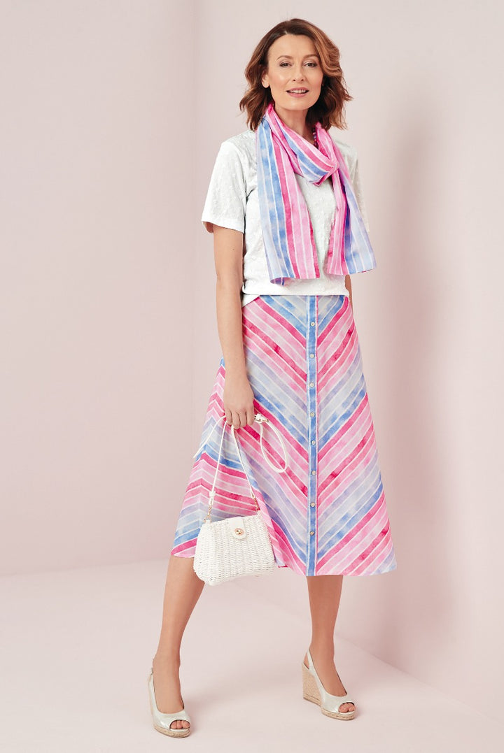 Lily Ella Collection summer attire with model wearing pastel pink and blue striped midi skirt, white textured t-shirt, matching striped scarf, and accessorized with white woven shoulder bag and wedge sandals.