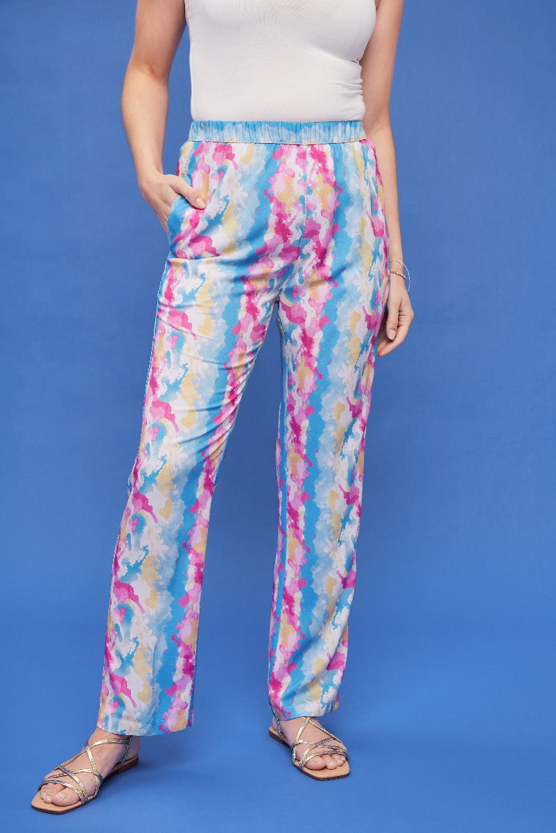 Lily Ella Collection vibrant multi-color floral print trousers, stylish women's casual wear, comfortable elastic waistband pants, paired with white top and metallic strap sandals on blue background.