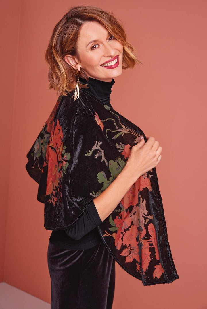Lily Ella Collection elegant black velvet floral-patterned kimono on a smiling model with a stylish turtleneck, complemented by chic earrings, poised against a soft pink background.