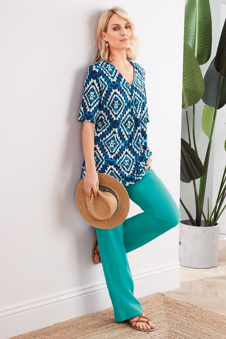 Lily Ella Collection stylish blue and white patterned tunic top with teal trousers, accessorized with tan woven hat and sandals, contemporary women's fashion outfit.