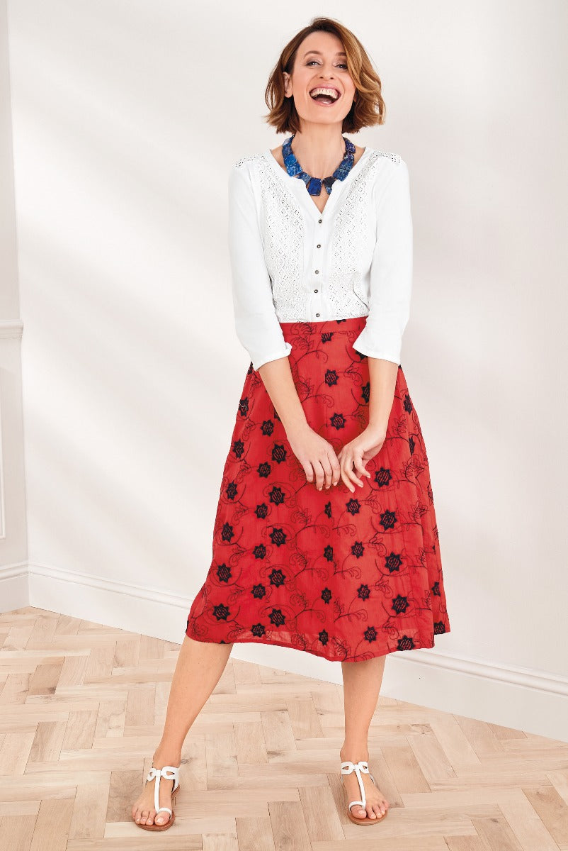Lily Ella Collection elegant woman modeling red floral skirt and white lace cardigan with statement blue necklace and white sandals, showcasing contemporary spring/summer women's fashion.