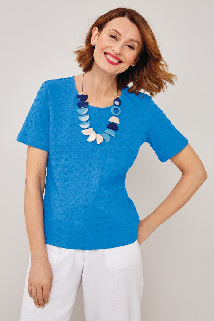 Lily Ella Collection vibrant blue lace-patterned top styled with contemporary white trousers and statement necklace on smiling model.