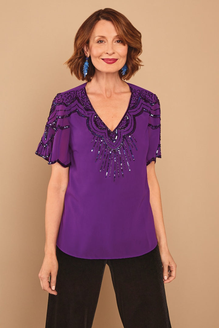Lily Ella Collection purple embellished tunic top with sequin and bead detailing, elegant flutter sleeves, and sophisticated v-neckline style on model matching with black trousers and statement blue earrings.
